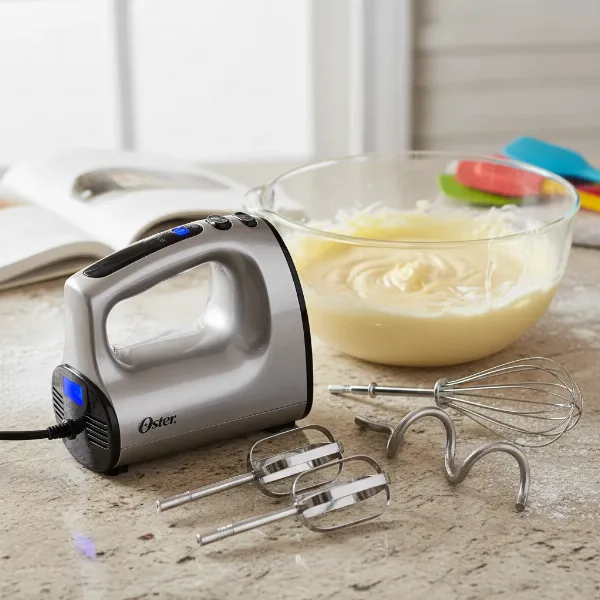 Oster HeatSoft hand mixer with various attachments (beaters, whisk, dough hooks) next to a bowl of batter.
